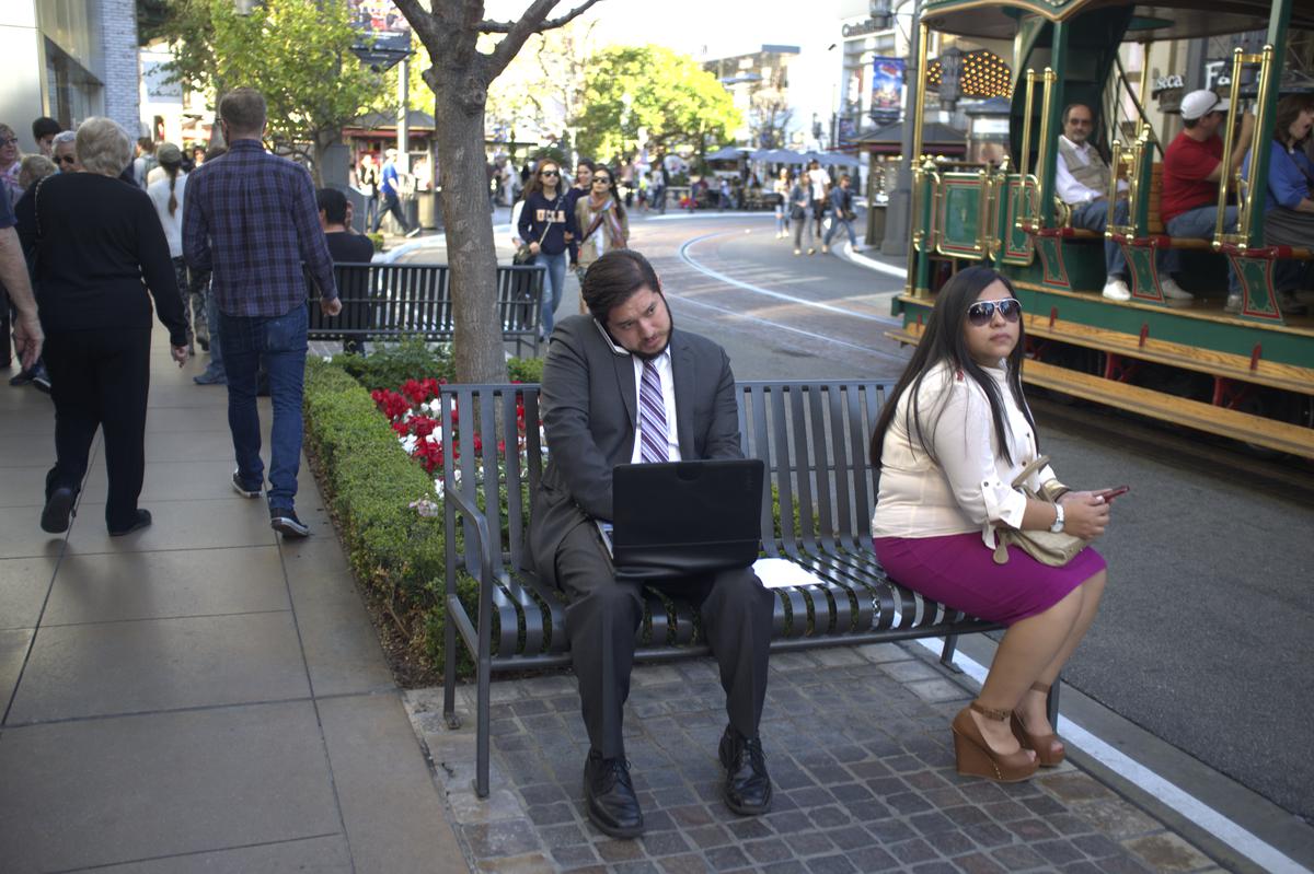 Man on his phone while using his laptop sharing a bench with a woman at an outdoor mall